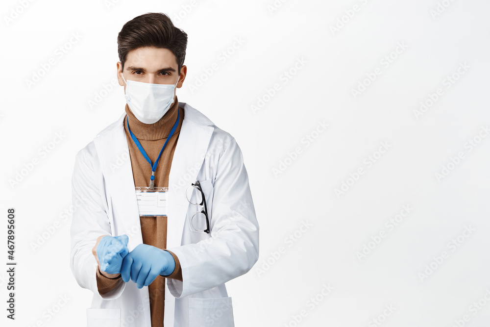 Covid 19. Doctor in medical face mask, put on gloves for patient examination, wearing clinic uniform, standing over white background Covid 19. Doctor in medical face mask, put on gloves for patient examination, wearing clinic uniform, standing over white background