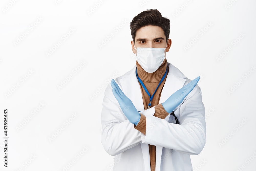Young doctor in medical face mask from covid 19, rubber gloves, making cross stop gesture, prohibit action, standing over white background Young doctor in medical face mask from covid 19, rubber gloves, making cross stop gesture, prohibit action, standing over white background