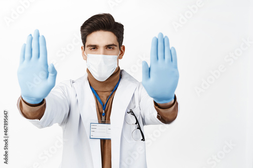 Portrait of mal doctor in face mask and robe, extend hands in gloves, showing stop gesture, taboo disapproval sign, standing over white background