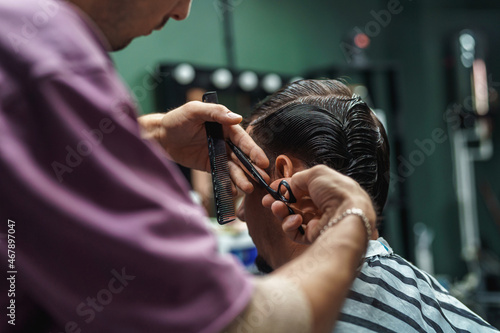 young barber guy gives a haircut to a bearded man sitting in a chair in a barbershop. A man cuts his hair with scissors and a comb. Men's haircuts and beard shaving. Haircut and edging of eyebrows.