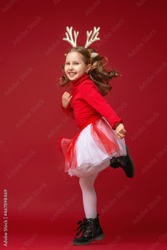 happy little girl in reindeer antlers running and jumping on red background