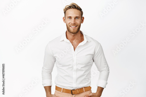 Handsome macho man with blond hair and beard, smiling and looking confident at camera, wearing white shirt, standing against studio background