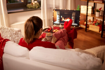 christmas, winter holidays and leisure concept - young woman watching tv with fireplace on screen and holding mug of marshmallow and whipped cream resting feet on table at cozy home