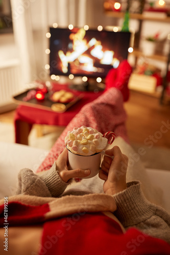 christmas, winter holidays and leisure concept - close up of young woman watching tv with fireplace on screen and holding mug of marshmallow and whipped cream resting feet on table at cozy home