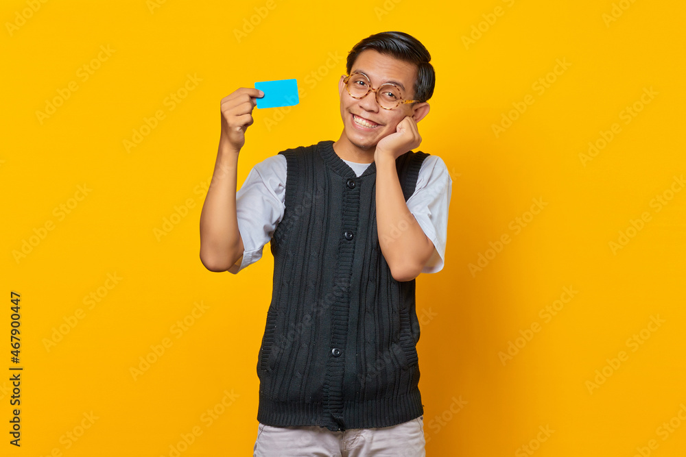 Smiling asian man holding credit card with open mouth and hands on cheeks on yellow background Smiling asian man holding credit card with open mouth and hands on cheeks on yellow background