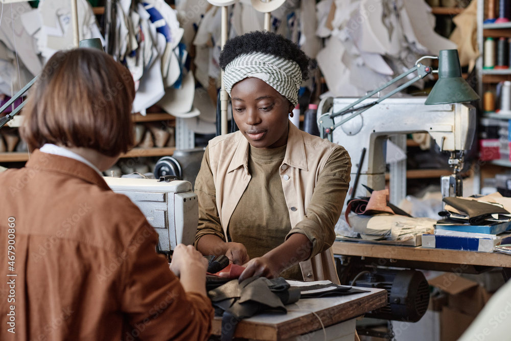 African young tailor sewing clothes from leather together with her colleague while they sitting at the table and working on sewing machine