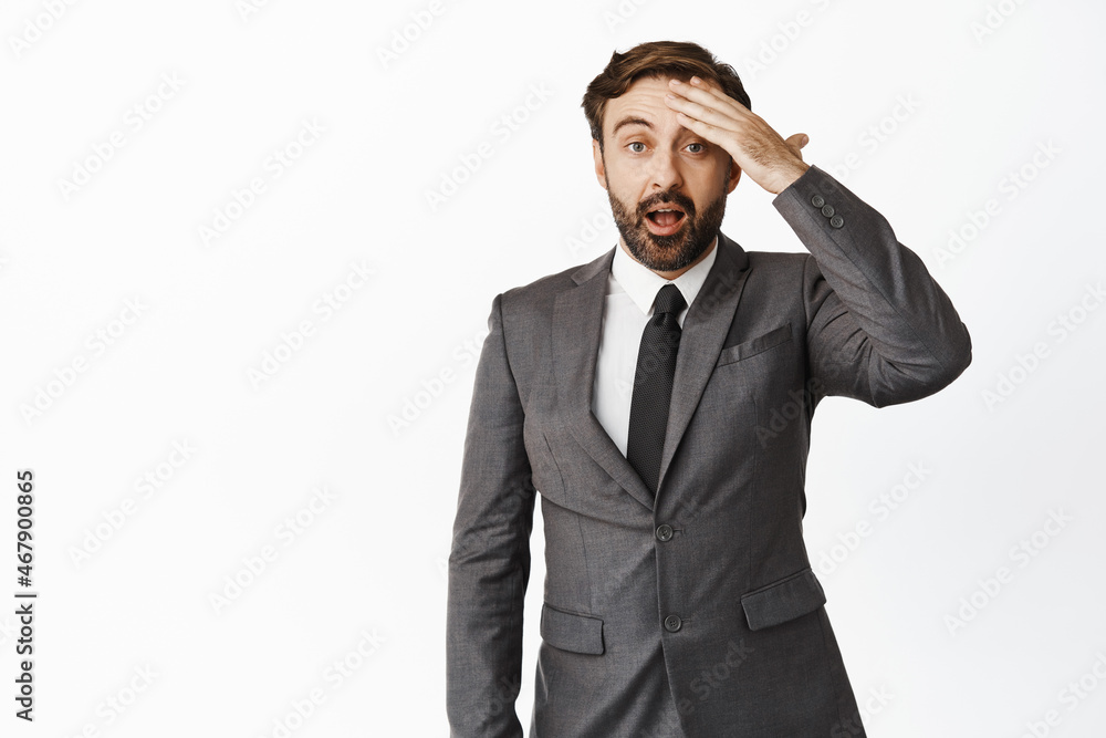 Shocked businessman in suit, touching forehead and looking forgetful, remember something, standing against white background Shocked businessman in suit, touching forehead and looking forgetful, remember something, standing against white background