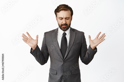 Confused businessman in suit raising empty hands up, looking down shocked, drop something below, standing over white background