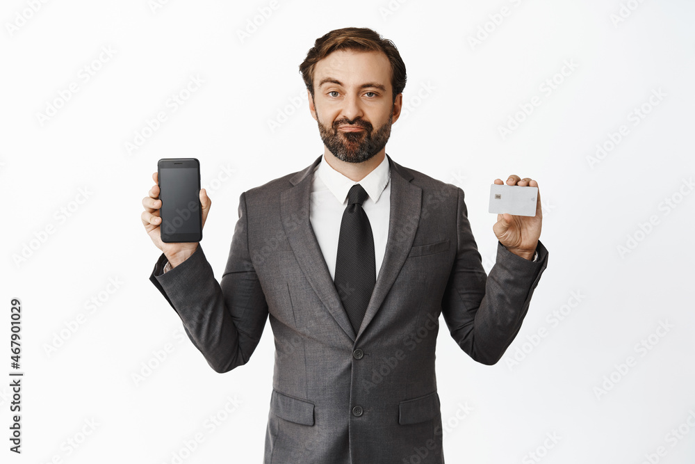 Skeptical man in suit shows his credit card and smartphone screen, smirk and look disappointed, negative opinion about phone app, white background Skeptical man in suit shows his credit card and smartphone screen, smirk and look disappointed, negative opinion about phone app, white background