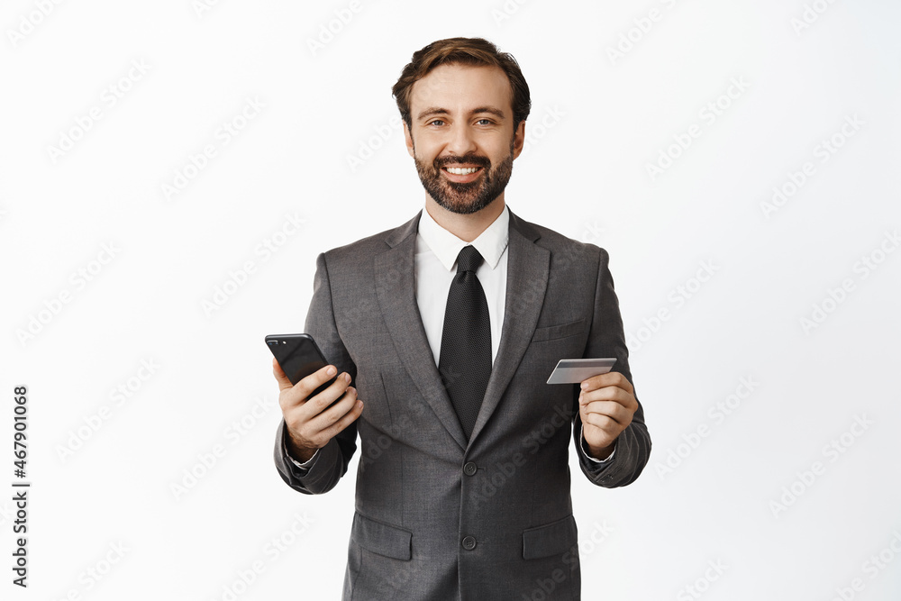 Business and technology concept. Handsome corporate man smiling pleased, holding credit card and smartphone, white background Business and technology concept. Handsome corporate man smiling pleased, holding credit card and smartphone, white background