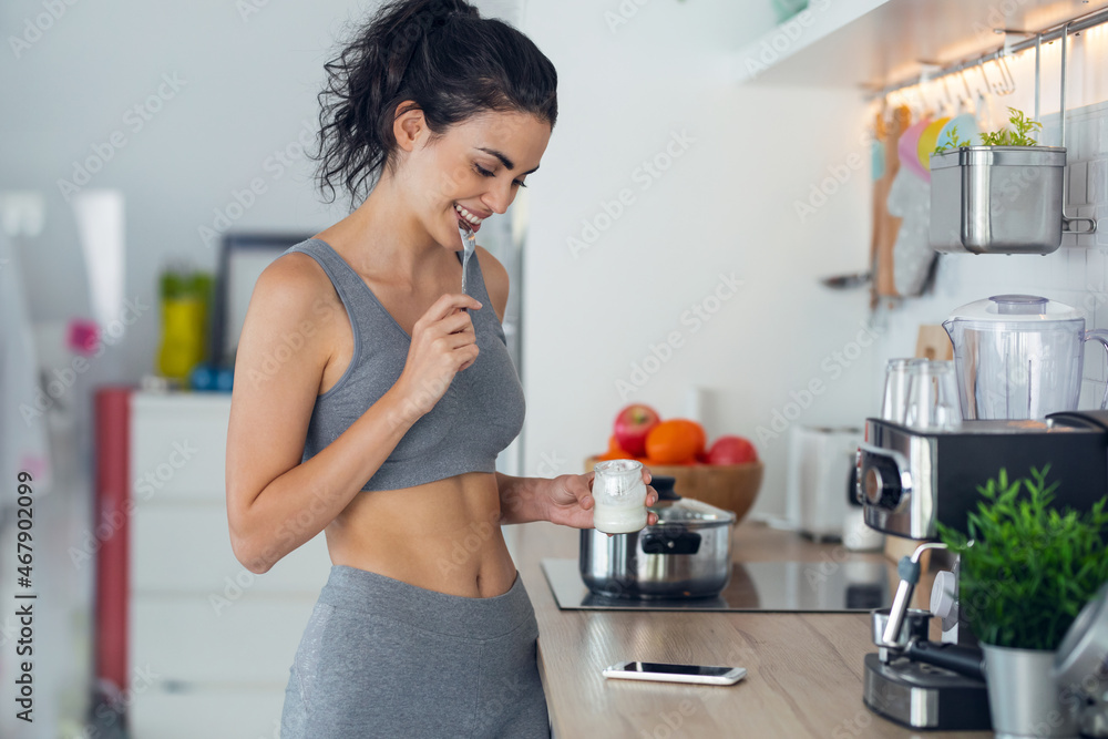 Fototapeta premium Sporty young sporty woman eating iogurt while using mobile phone standing in the kitchen at home.