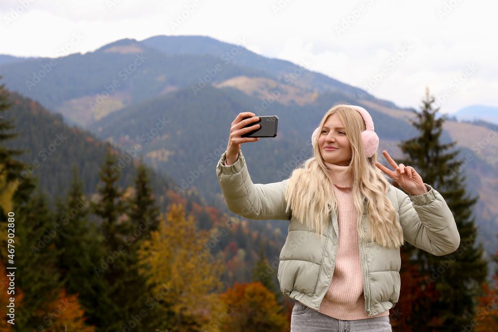 Young woman in warm earmuffs taking selfie on autumn day