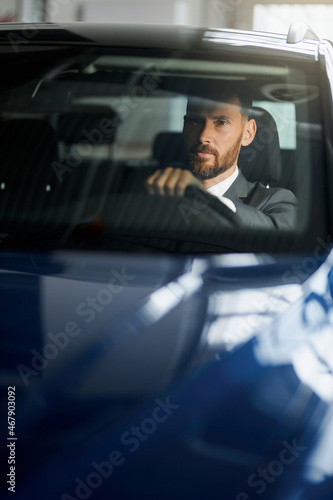 Successful man in stylish suit examining cars at showroom. Bearded male customer sitting in side blue auto. Businessman making expensive purchase.
