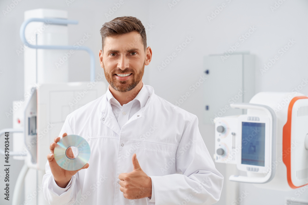 Handsome brunette doctor showing finger up, no illness result after ultrasound examine. Professional smiling radiologist looking at camera holding silver CD plate with information about procedure. Handsome brunette doctor showing finger up, no illness result after ultrasound examine. Professional smiling radiologist looking at camera holding silver CD plate with information about procedure.