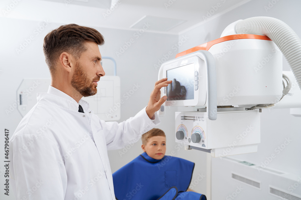 Side view of brunette doctor with beard touching screen of usg modern tool, starting procedure of diagnostic. Professional radiologist doing ultrasound diagnostic for little boy wearing blue robe. Side view of brunette doctor with beard touching screen of usg modern tool, starting procedure of diagnostic. Professional radiologist doing ultrasound diagnostic for little boy wearing blue robe.