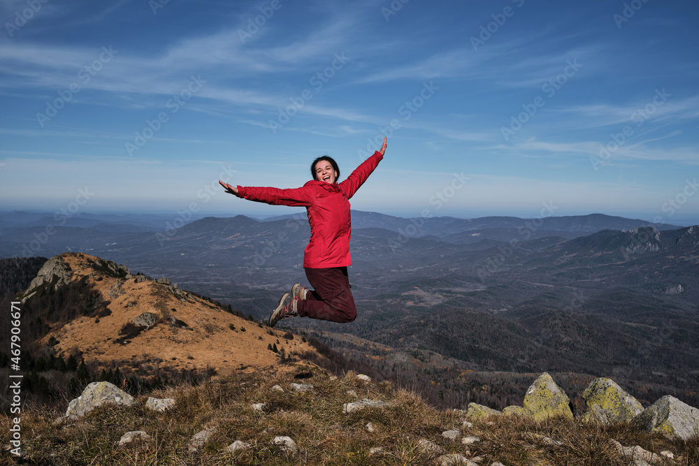 Enjoy adventure. Female traveler in bright red jacket jumps merrily on top of mountain against background of autumn landscape. Hairstyle of dreadlocks of tourist girl. Enjoy adventure. Female traveler in bright red jacket jumps merrily on top of mountain against background of autumn landscape. Hairstyle of dreadlocks of tourist girl.