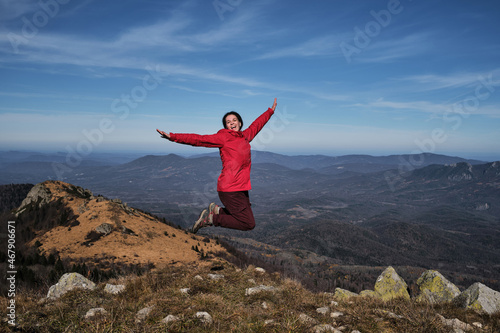 Enjoy adventure. Female traveler in bright red jacket jumps merrily on top of mountain against background of autumn landscape. Hairstyle of dreadlocks of tourist girl.