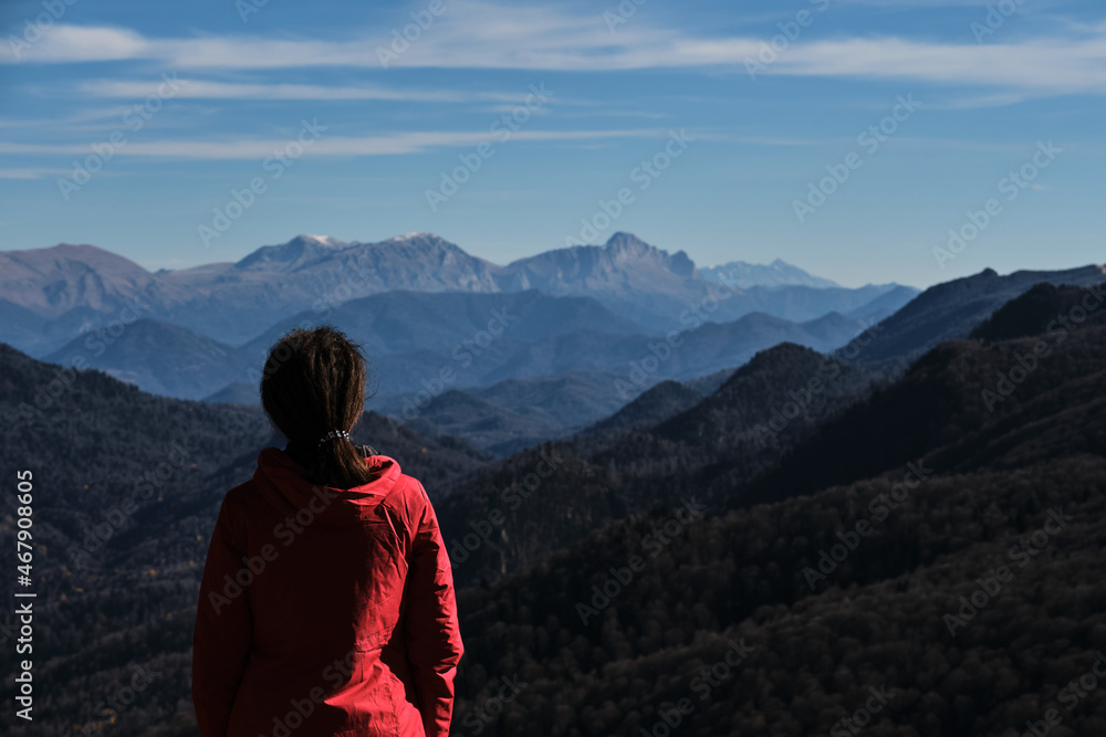 Hairstyle of dreadlocks of tourist girl. Female traveler in bright red jacket stands with her back turned and looks at great snow capped mountains on horizon. Hairstyle of dreadlocks of tourist girl. Female traveler in bright red jacket stands with her back turned and looks at great snow capped mountains on horizon.