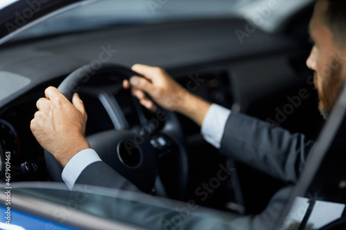 Side view of bearded businessman in stylish suit holding hands on steering wheel while sitting inside modern car. Male client buying new auto at salon.
