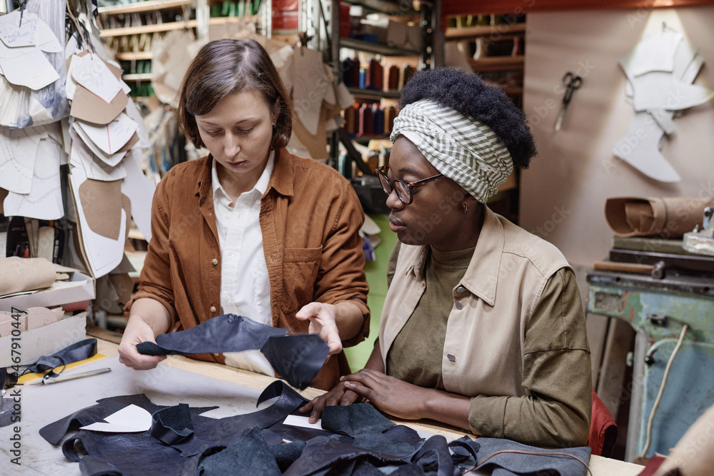 Two tailors sitting at their workplaces and sewing pieces of the fabric in team during their work in the workshop