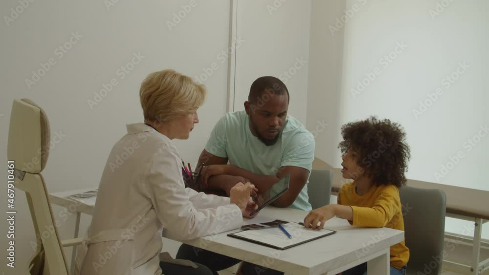 Pretty middle aged female pediatrician writing digital prescription on tablet pc, explaining treatment plan of sick cute elementary age african american boy to worried father in medical office. Pretty middle aged female pediatrician writing digital prescription on tablet pc, explaining treatment plan of sick cute elementary age african american boy to worried father in medical office.
