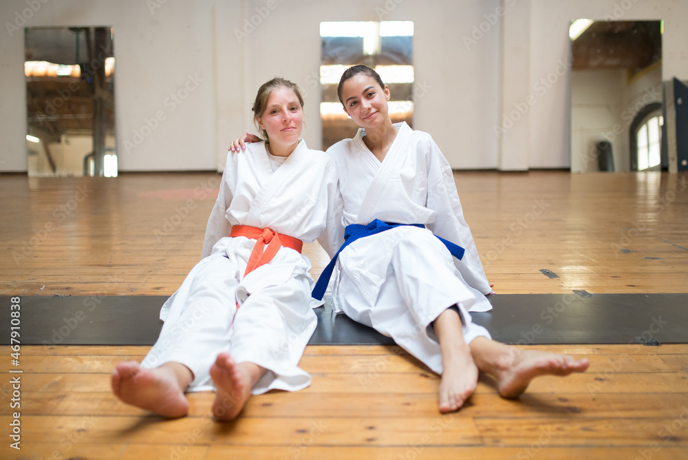 Happy young female karatekas sitting and hugging together. Cheerful Caucasian girls resting after karate class in studio. Sports, martial arts, healthy lifestyle concept