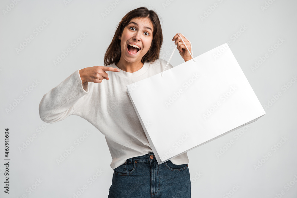 Cheerful positive woman pointing finger at shopping bag in her hand. Happy shopping and black Friday concept. Indoor studio shot isolated on white background