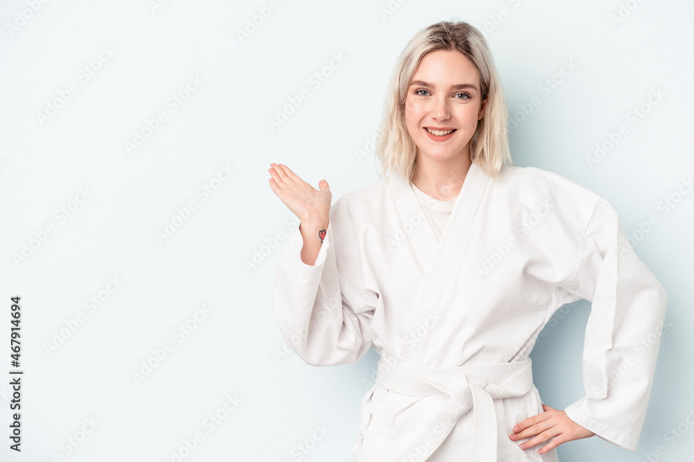 Young caucasian woman doing karate isolated on blue background showing a copy space on a palm and holding another hand on waist. Young caucasian woman doing karate isolated on blue background showing a copy space on a palm and holding another hand on waist.