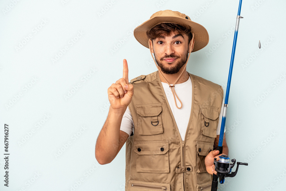 Young mixed race fisherman holding a rod isolated on blue background showing number one with finger. Young mixed race fisherman holding a rod isolated on blue background showing number one with finger.