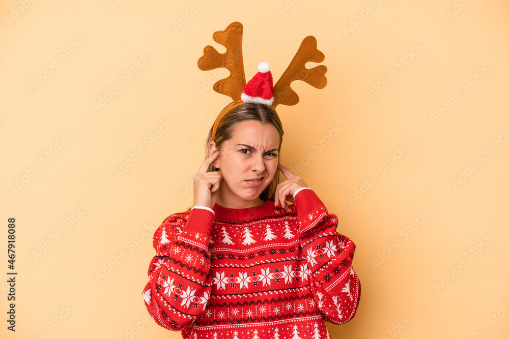 Young caucasian woman wearing a christmas reindeer hat isolated on yellow background covering ears with hands. Young caucasian woman wearing a christmas reindeer hat isolated on yellow background covering ears with hands.