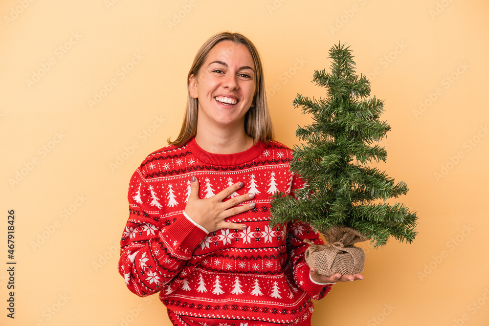 Young caucasian woman holding a little christmas tree isolated on yellow background laughs out loudly keeping hand on chest. Young caucasian woman holding a little christmas tree isolated on yellow background laughs out loudly keeping hand on chest.