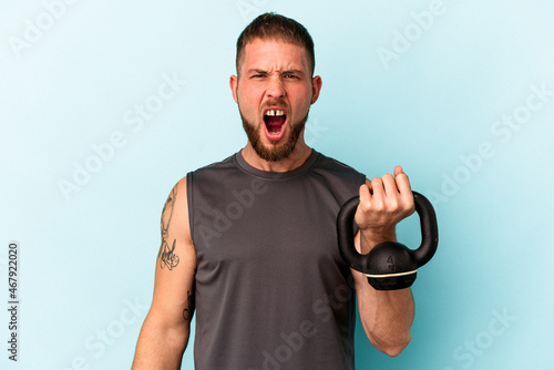 Young caucasian man holding ketlebell isolated on blue background screaming very angry and aggressive. photo