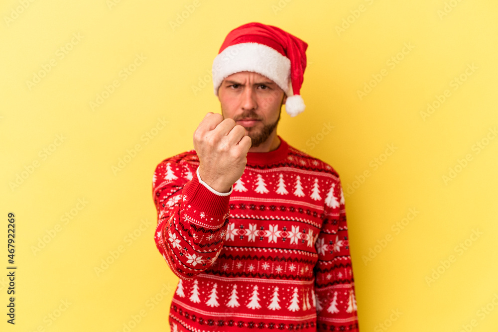 Young caucasian man celebrating Christmas isolated on yellow background showing fist to camera, aggressive facial expression. Young caucasian man celebrating Christmas isolated on yellow background showing fist to camera, aggressive facial expression.