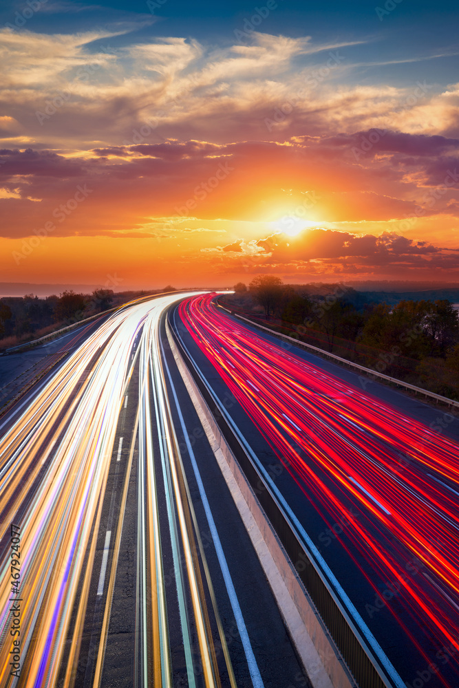 Trails of cars lights on the asphalt car road. Sunset time with clouds and sun. Drive forward! Transport creative background. Long exposure, motion and blur.