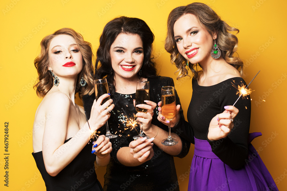 three young women with a champagne glasses and sparklers over yellow background