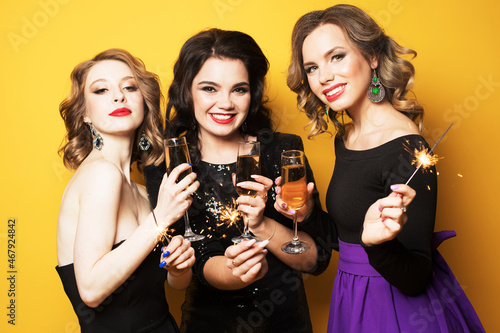 three young women with a champagne glasses and sparklers over yellow background