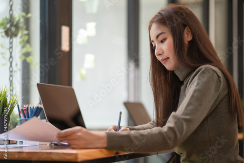Businesswoman writing a note while using laptop at a table in an office.