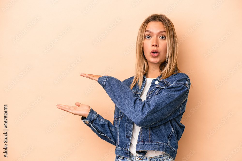 Young caucasian woman isolated on beige background shocked and amazed holding a copy space between hands. Young caucasian woman isolated on beige background shocked and amazed holding a copy space between hands.