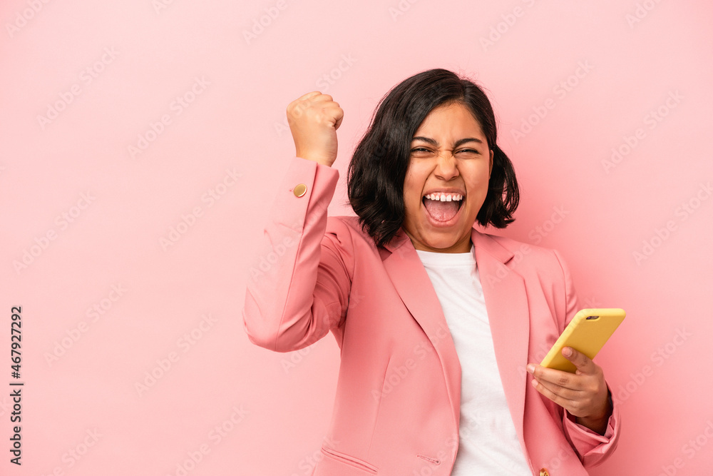Young latin woman holding mobile phone isolated on pink background raising fist after a victory, winner concept. Young latin woman holding mobile phone isolated on pink background raising fist after a victory, winner concept.