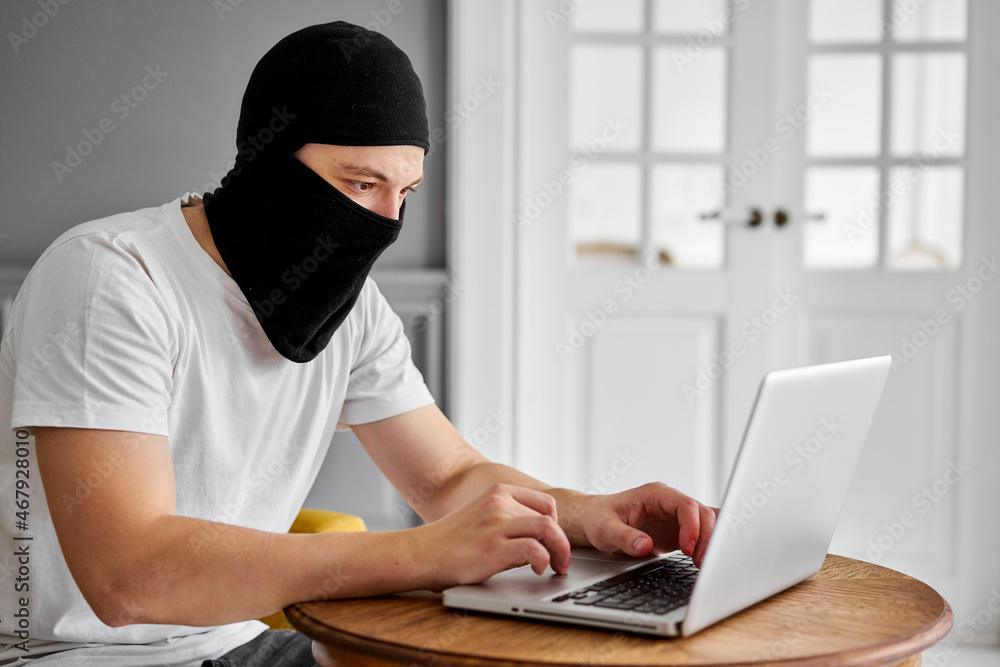 portrait of unrecognizable young hacker in black mask using laptop. Concept of cyber security and data protection portrait of unrecognizable young hacker in black mask using laptop. Concept of cyber security and data protection