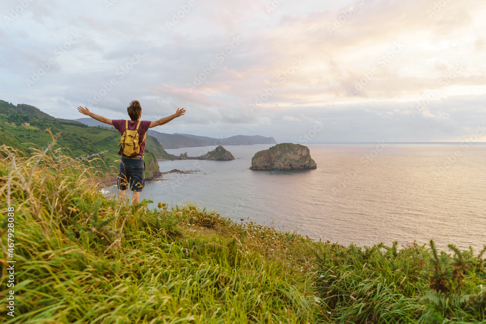 Panoramic view of unrecognizable man with arms wide open at sunset in a cliff. Horizontal view of backpacker traveling outdoors with the blue ocean on the background.