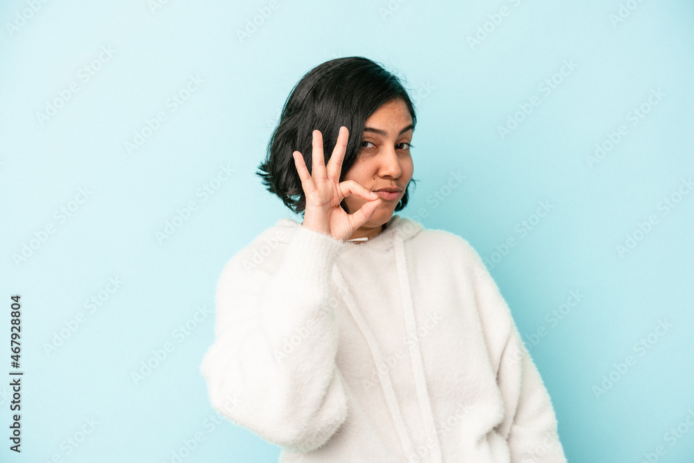 Young latin woman isolated on blue background winks an eye and holds an okay gesture with hand. Young latin woman isolated on blue background winks an eye and holds an okay gesture with hand.