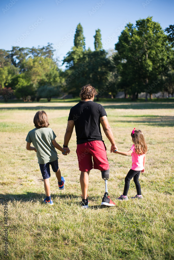 Fototapeta premium Back view of happy father with disability walking with children. Man with mechanical leg having walk in park with boy and girl. Disability, family, love concept