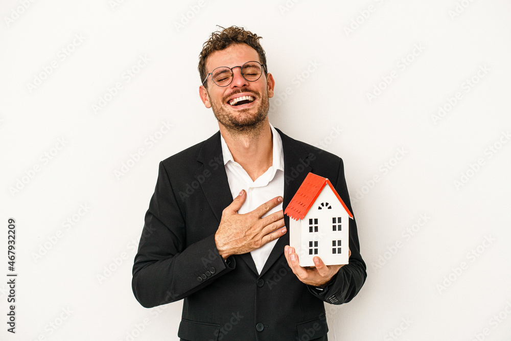 Young caucasian real estate holding a toy home isolated on white background laughs out loudly keeping hand on chest. Young caucasian real estate holding a toy home isolated on white background laughs out loudly keeping hand on chest.