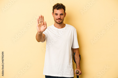 Young caucasian man holding crutch isolated on yellow background standing with outstretched hand showing stop sign, preventing you. photo