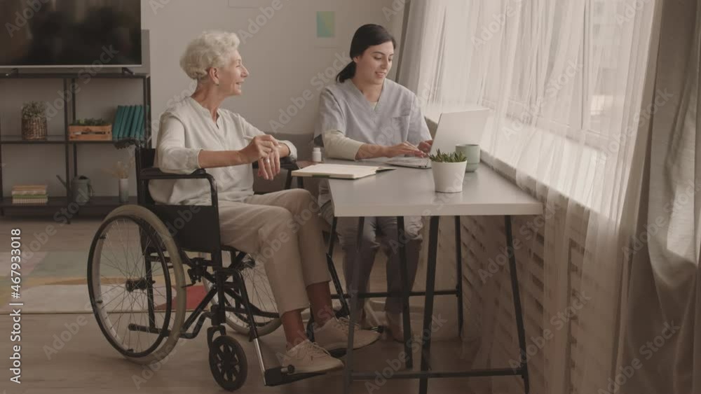 Stab full shot of young female social worker helping senior woman in wheelchair with making online payments on laptop, sitting by desk and having conversation Stab full shot of young female social worker helping senior woman in wheelchair with making online payments on laptop, sitting by desk and having conversation