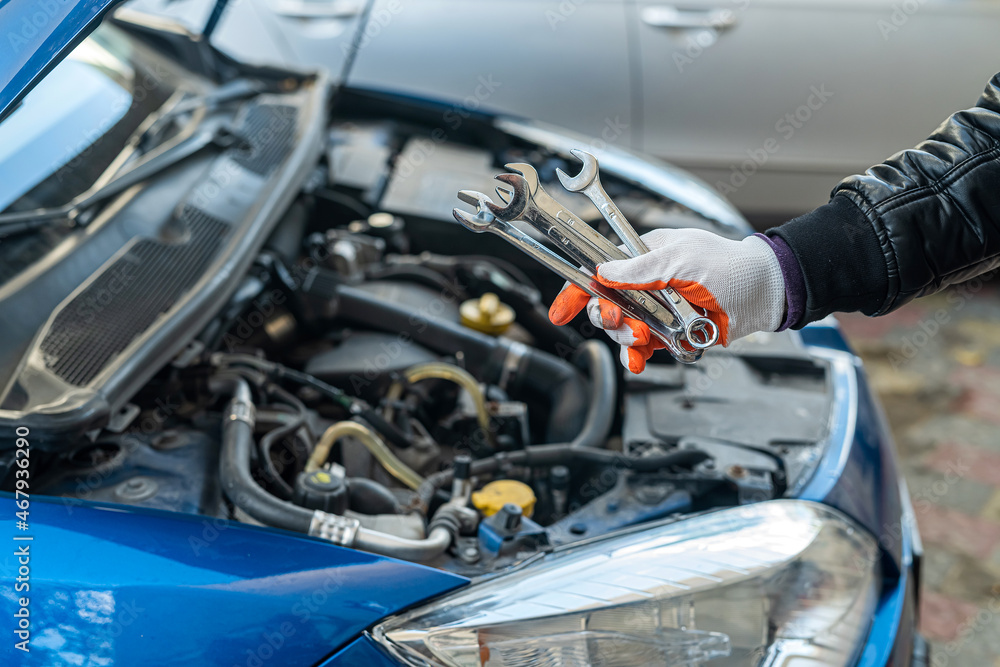 Man's gloved mechanic's hand holds car tools near a painted car with an open hood Man's gloved mechanic's hand holds car tools near a painted car with an open hood