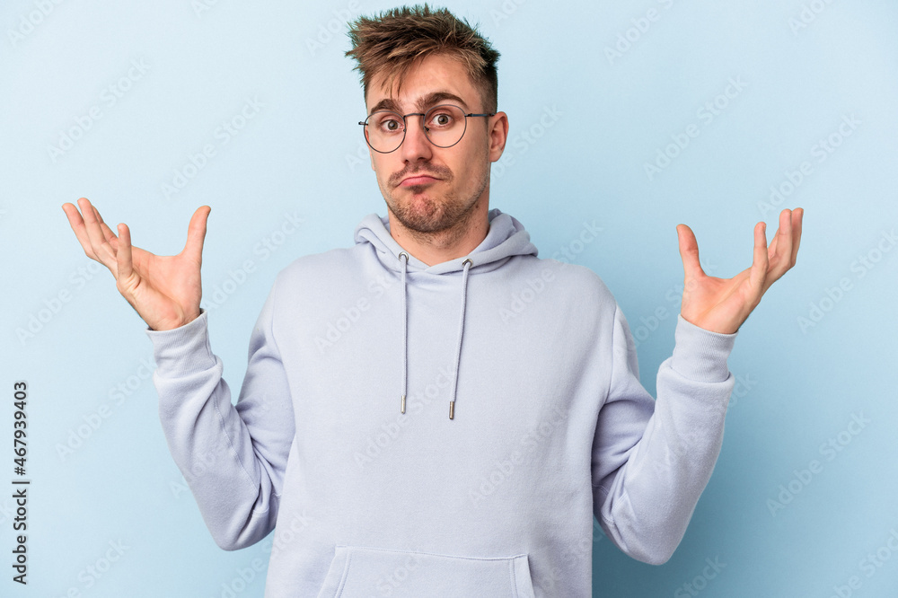 Young caucasian man isolated on blue background doubting and shrugging shoulders in questioning gesture. Young caucasian man isolated on blue background doubting and shrugging shoulders in questioning gesture.