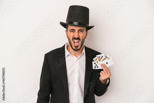 Young magician man holding a magic card isolated on white background screaming very angry and aggressive.