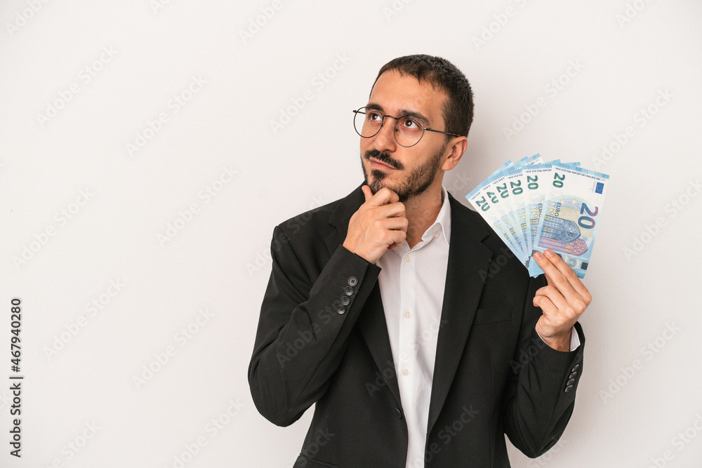 Young caucasian business man holding banknotes isolated on white background looking sideways with doubtful and skeptical expression. Young caucasian business man holding banknotes isolated on white background looking sideways with doubtful and skeptical expression.